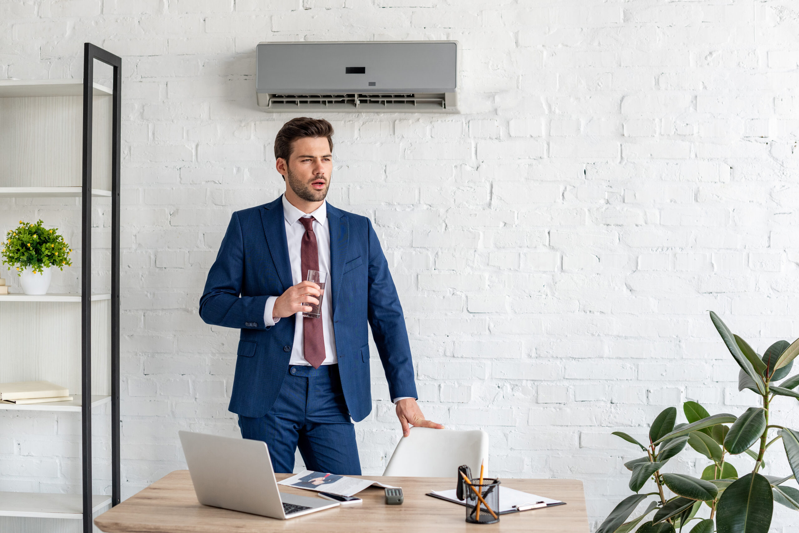 handsome businessman holding glass of water while standing near workplace under air conditioner